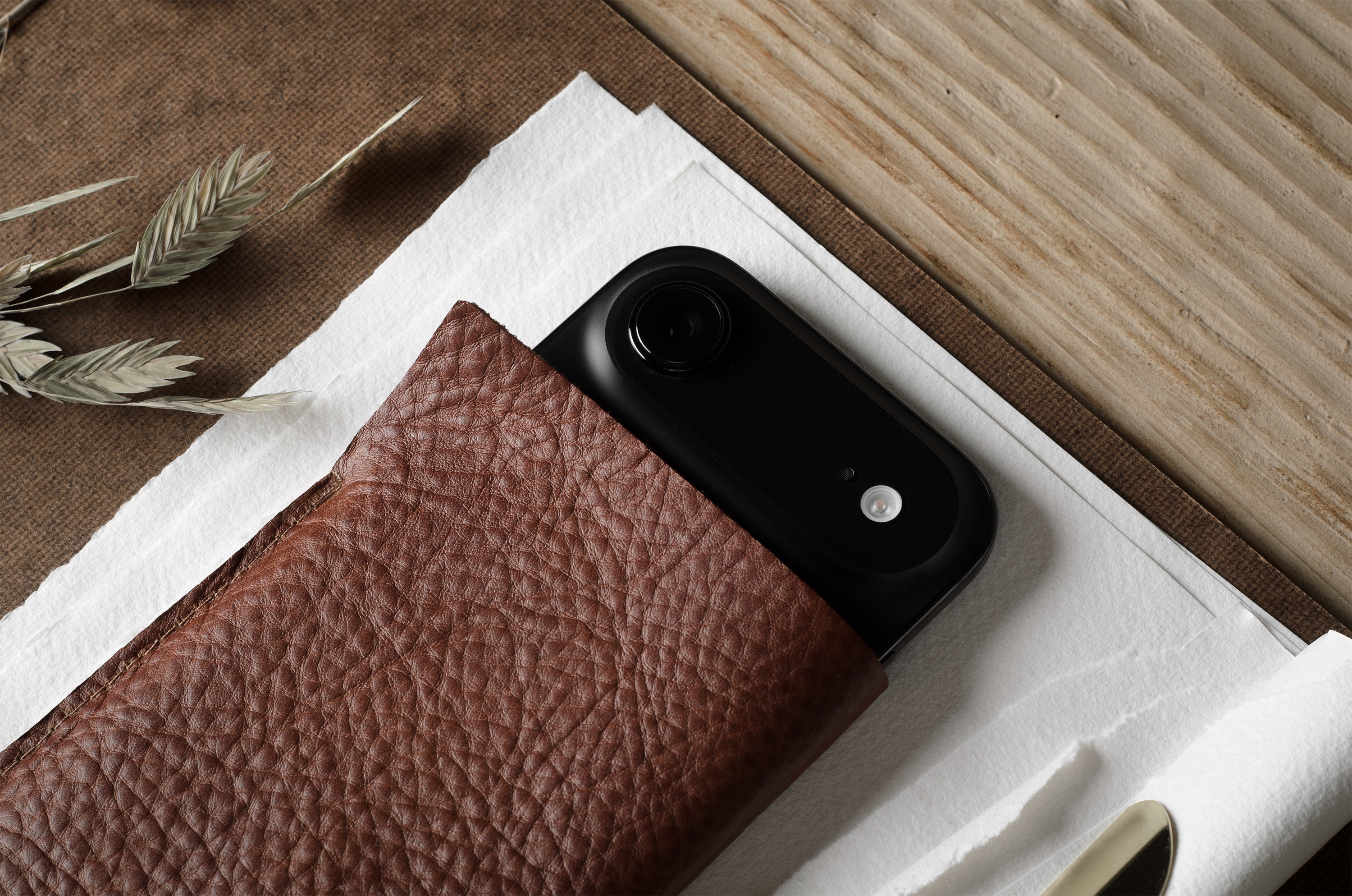 A close-up of a black smartphone partially tucked into a brown textured leather iphone case, placed on white paper over a wood-grain surface with dried grasses in the corner.