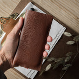 A hand holding a brown pebbled leather iphone case over a styled flat-lay of a brown notebook, white paper, dried grass, and a small white bead on a light wood surface.