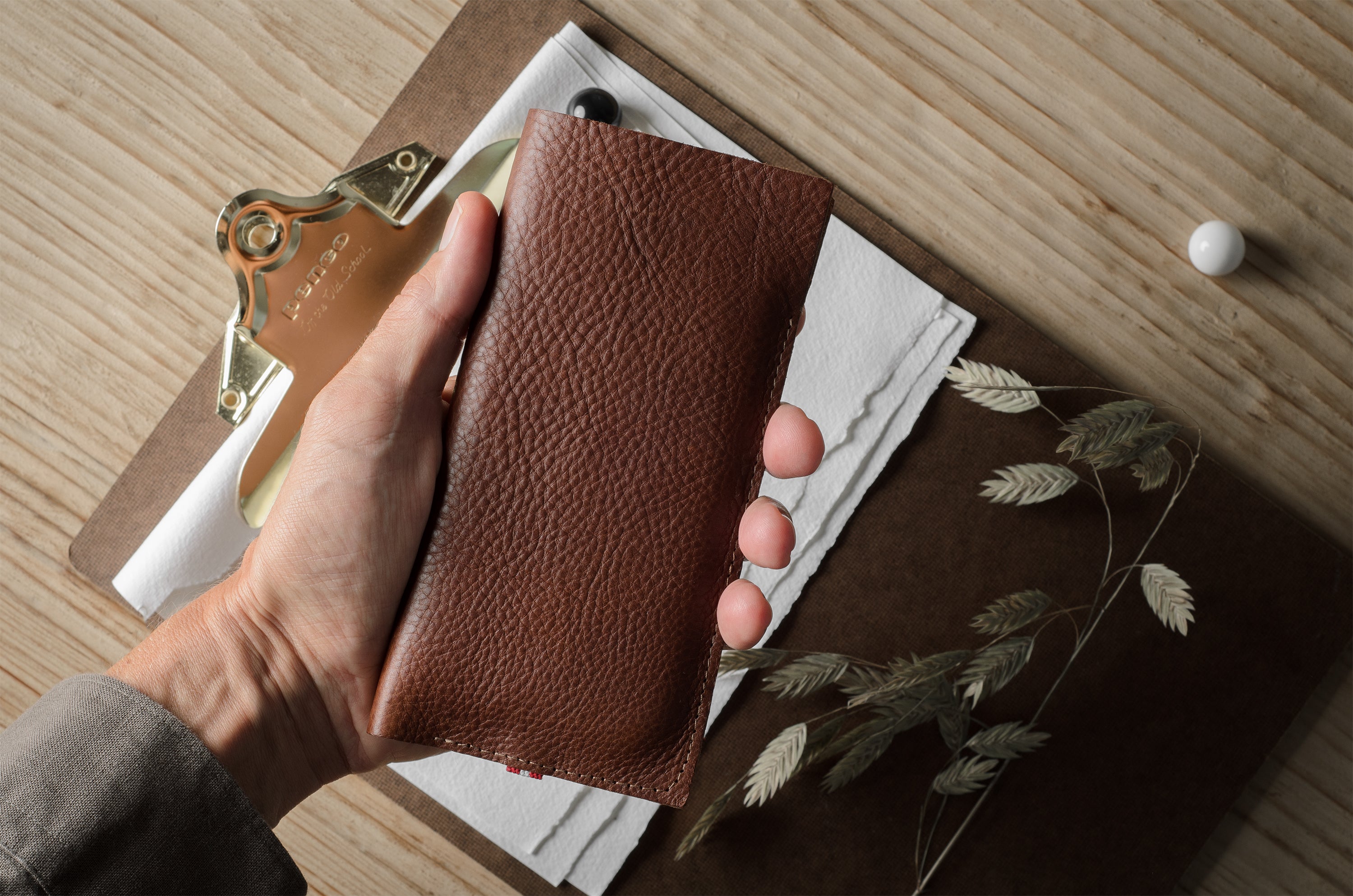 A hand holding a brown pebbled leather iphone case over a styled flat-lay of a brown notebook, white paper, dried grass, and a small white bead on a light wood surface.