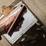 A hand holds a brown leather iphone case over a clipboard with white paper, on a light wood tabletop. A small white spherical object and dried botanical stems are nearby.