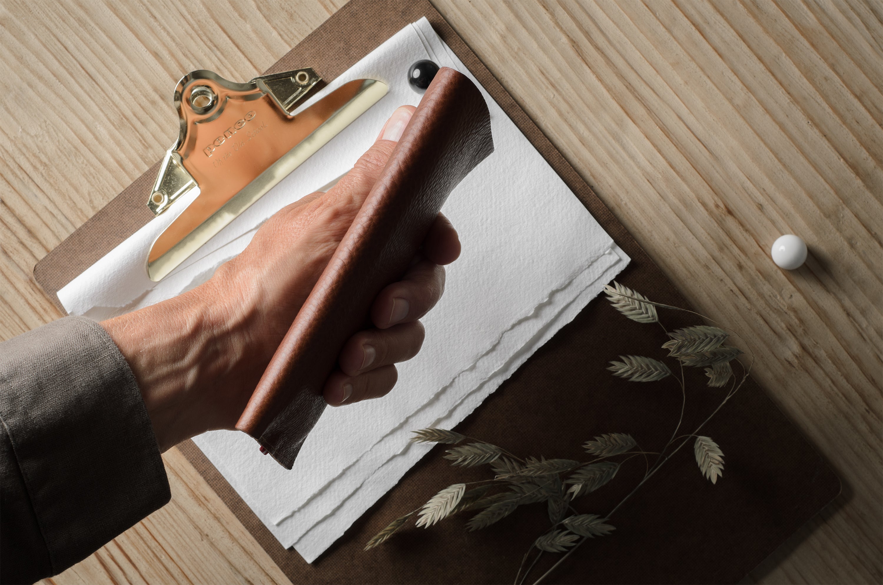 A hand holds a brown leather iphone case over a clipboard with white paper, on a light wood tabletop. A small white spherical object and dried botanical stems are nearby.