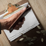 A close-up of a hand holding a brown iphone case over a clipboard with white paper, set on a light wood tabletop with dried botanicals nearby.
