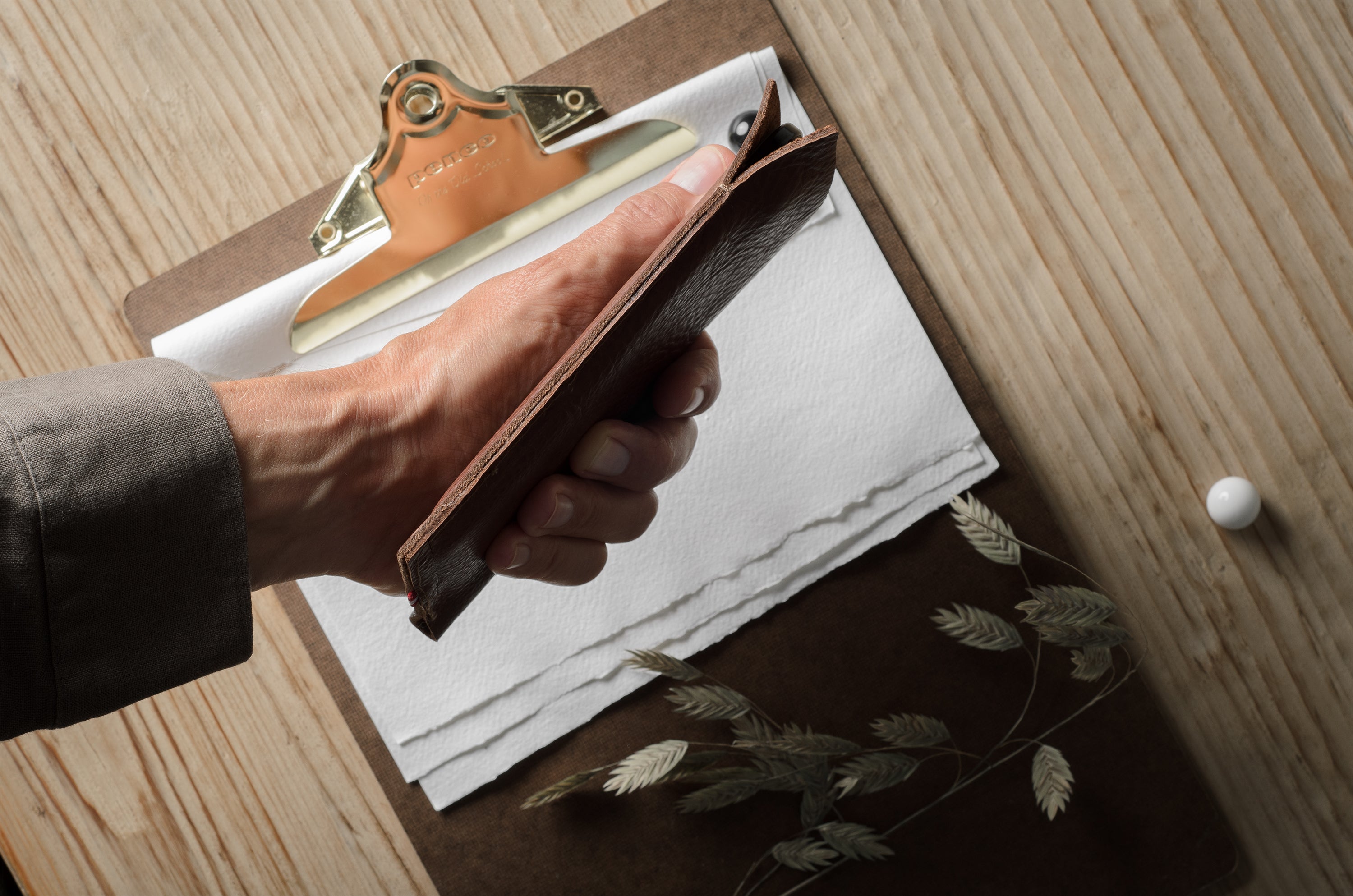 A close-up of a hand holding a brown iphone case over a clipboard with white paper, set on a light wood tabletop with dried botanicals nearby.