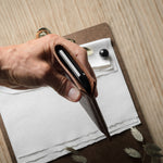 A close-up of a hand holding an open brown leather iphone case over a clipboard with papers on a light wood tabletop.