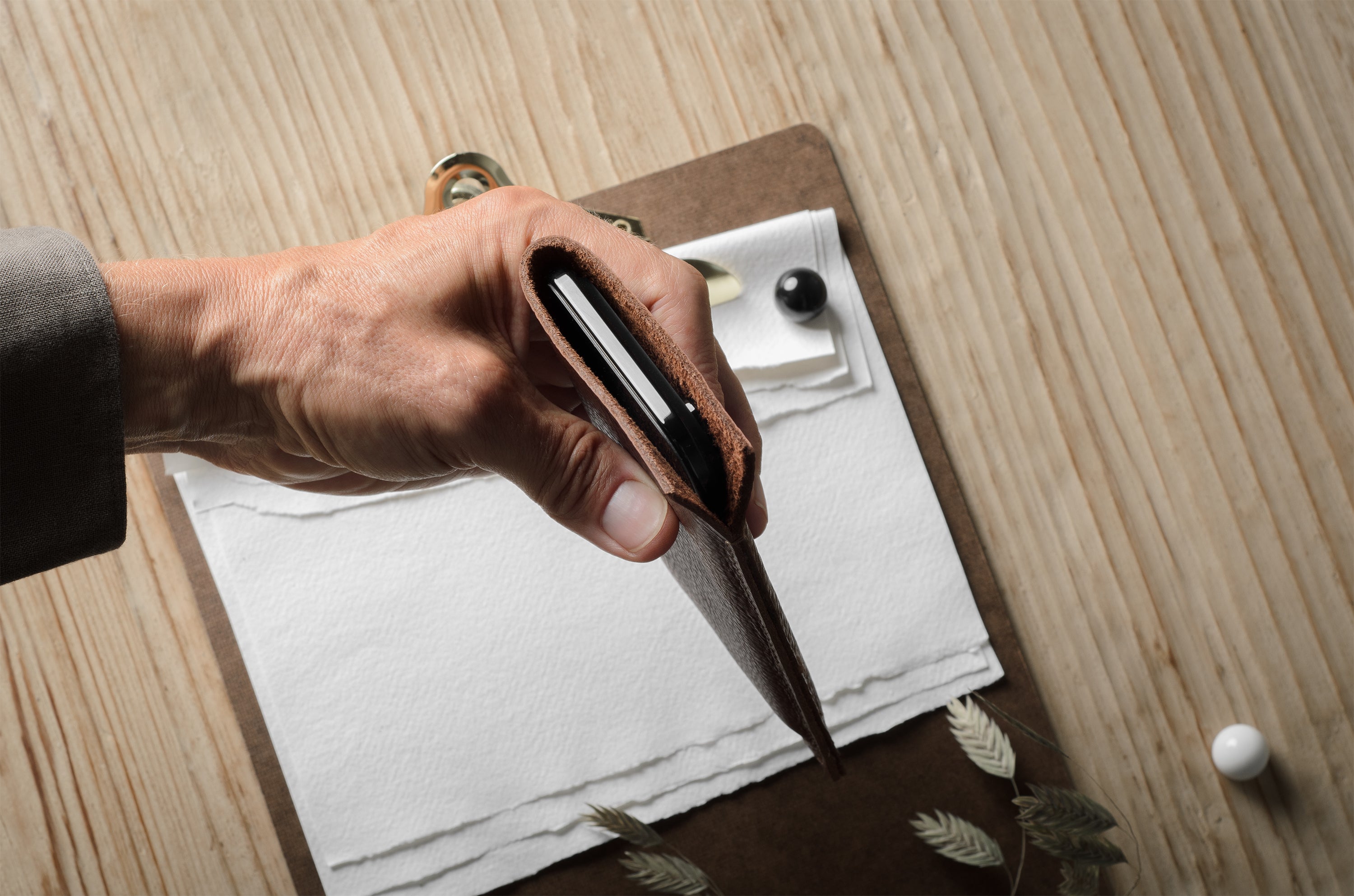 A close-up of a hand holding an open brown leather iphone case over a clipboard with papers on a light wood tabletop.