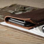 A dark brown textured leather iphone case holding a black smartphone, styled on a wooden board with dried wheat, a white napkin, and a black decorative object.