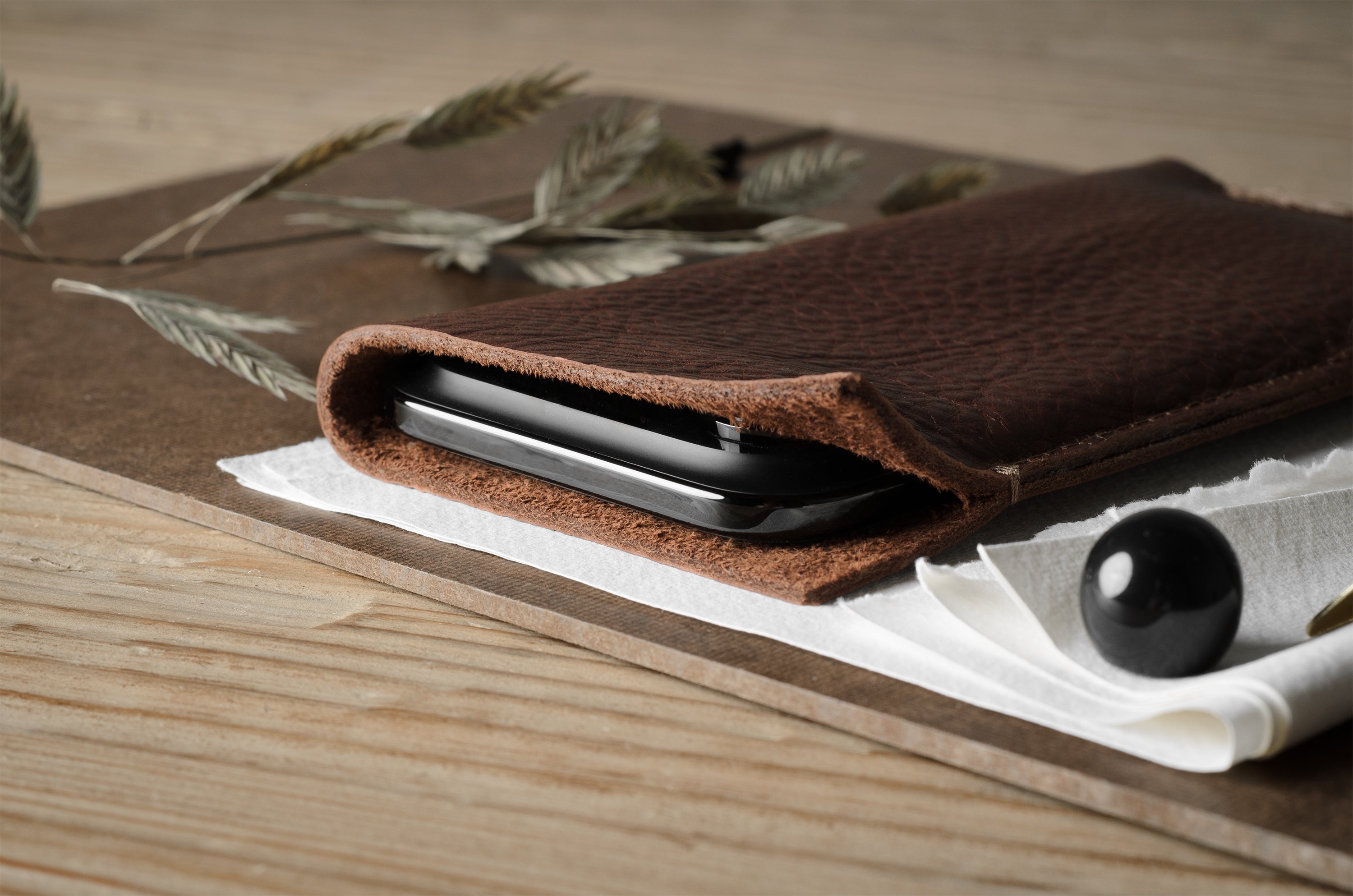 A dark brown textured leather iphone case holding a black smartphone, styled on a wooden board with dried wheat, a white napkin, and a black decorative object.