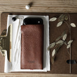 A brown pebbled leather iphone case holding a black smartphone, arranged on a clipboard with papers, a brass clip, dried grass stems, and a wooden tabletop background.