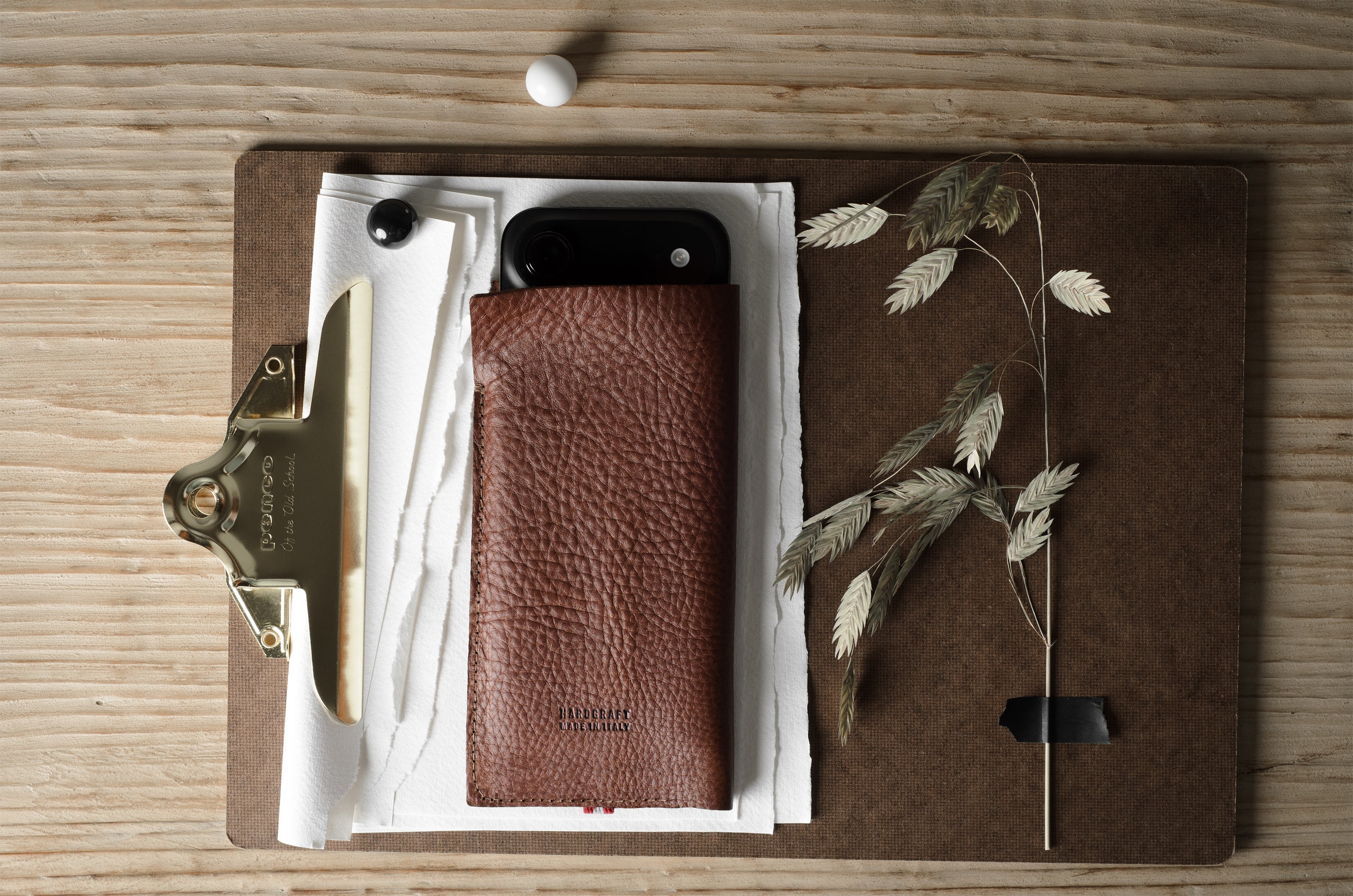 A brown pebbled leather iphone case holding a black smartphone, arranged on a clipboard with papers, a brass clip, dried grass stems, and a wooden tabletop background.