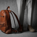 Brown leather backpack on a gray surface with a person wearing light-colored pants and shoes in the background.
