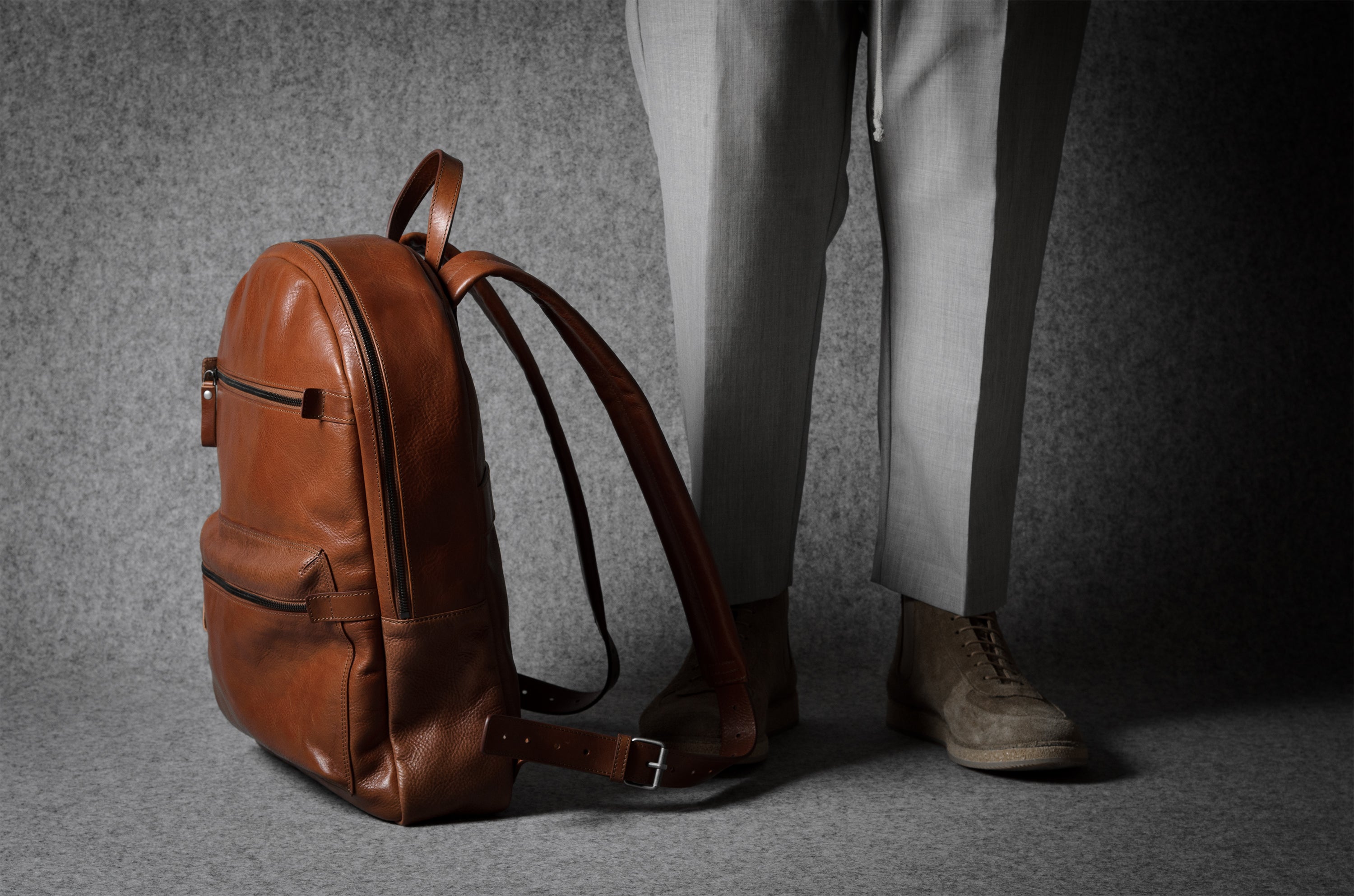 Brown leather backpack on a gray surface with a person wearing light-colored pants and shoes in the background.
