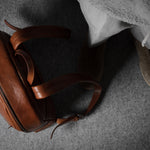 Brown leather backpack on a gray surface with a person wearing brown shoes.