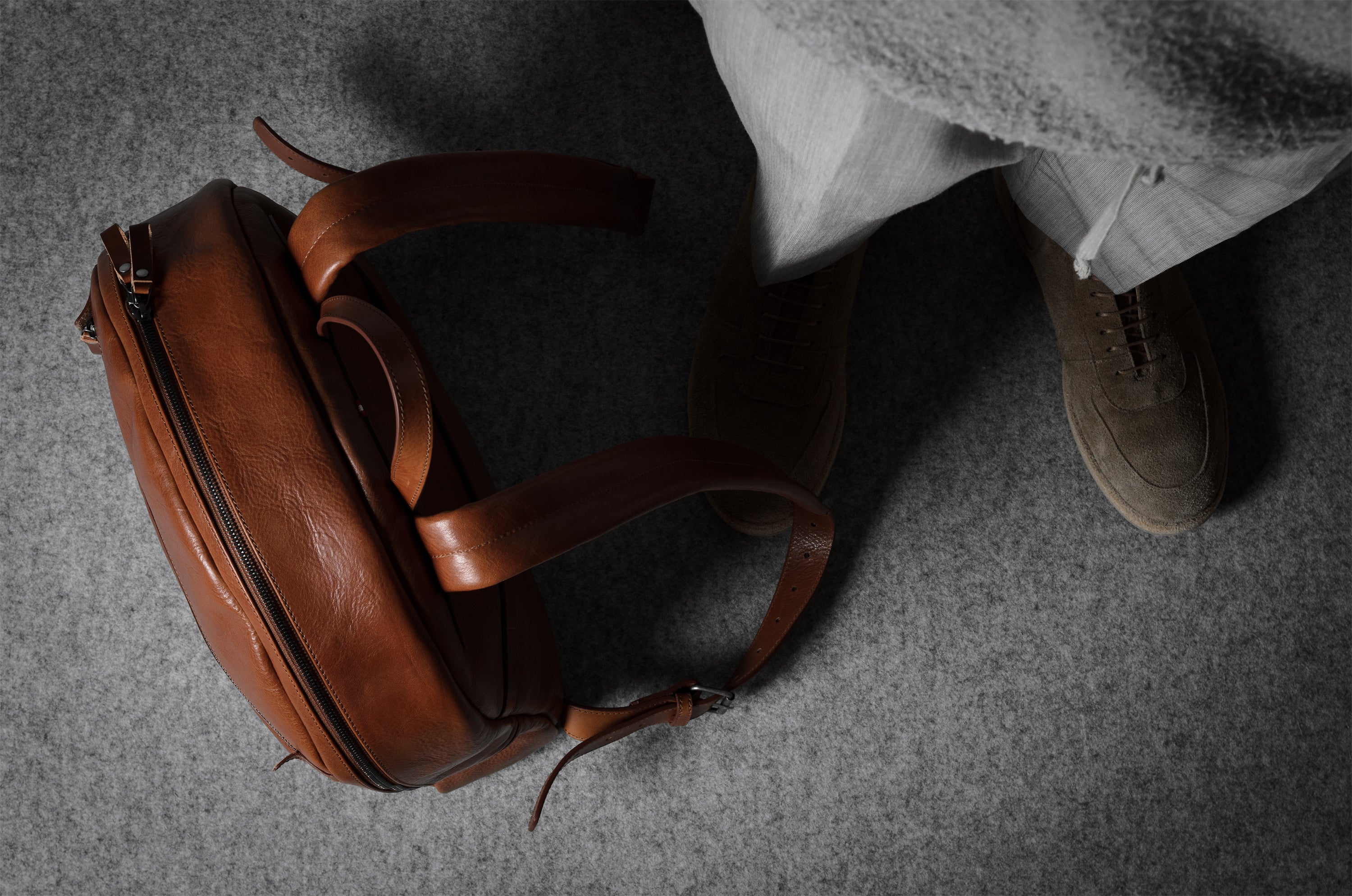 Brown leather backpack on a gray surface with a person wearing brown shoes.