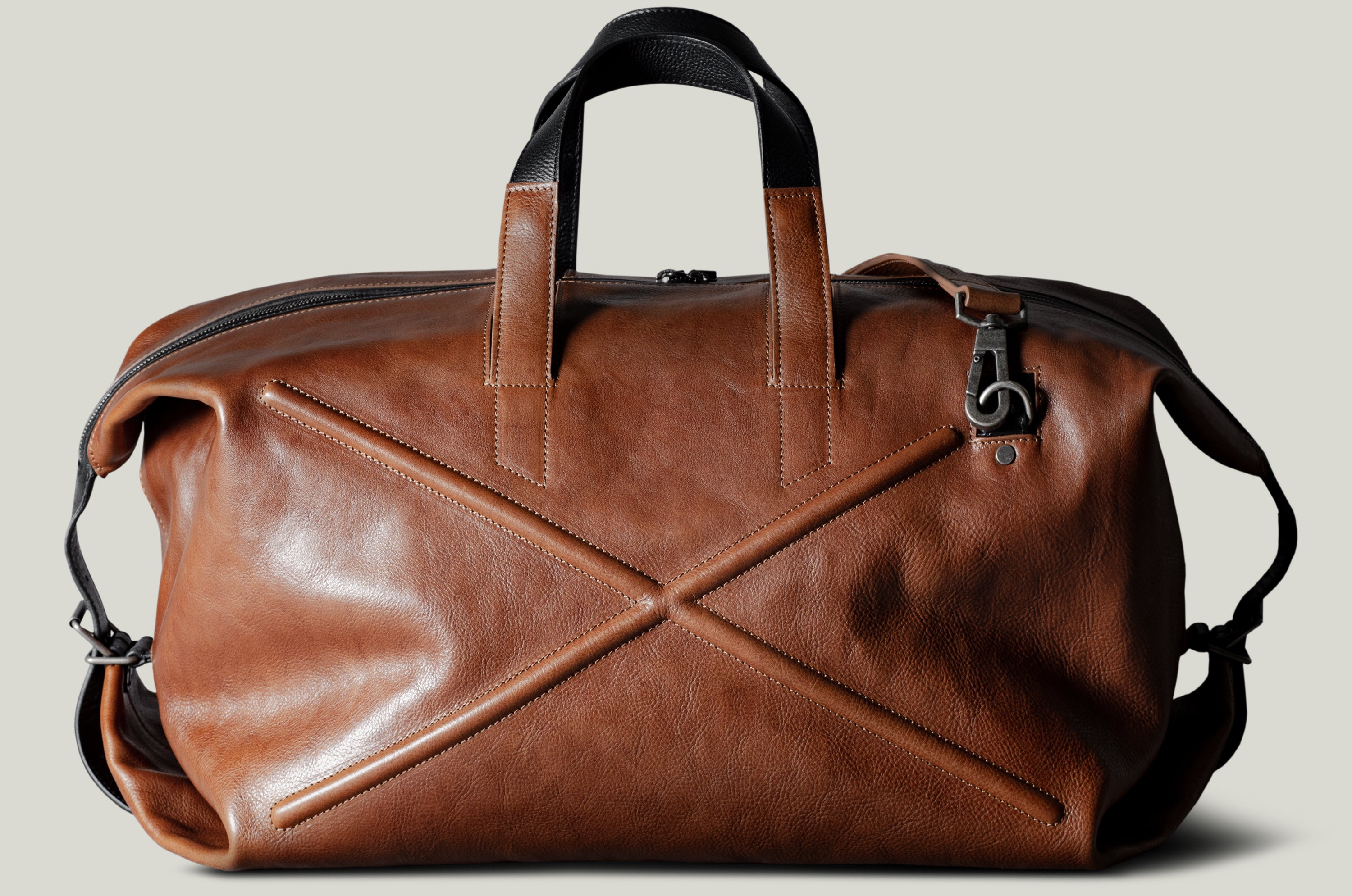 Brown leather duffel bag with black handles and strap on a light gray background