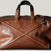 Brown leather duffel bag with black handles and strap on a light gray background