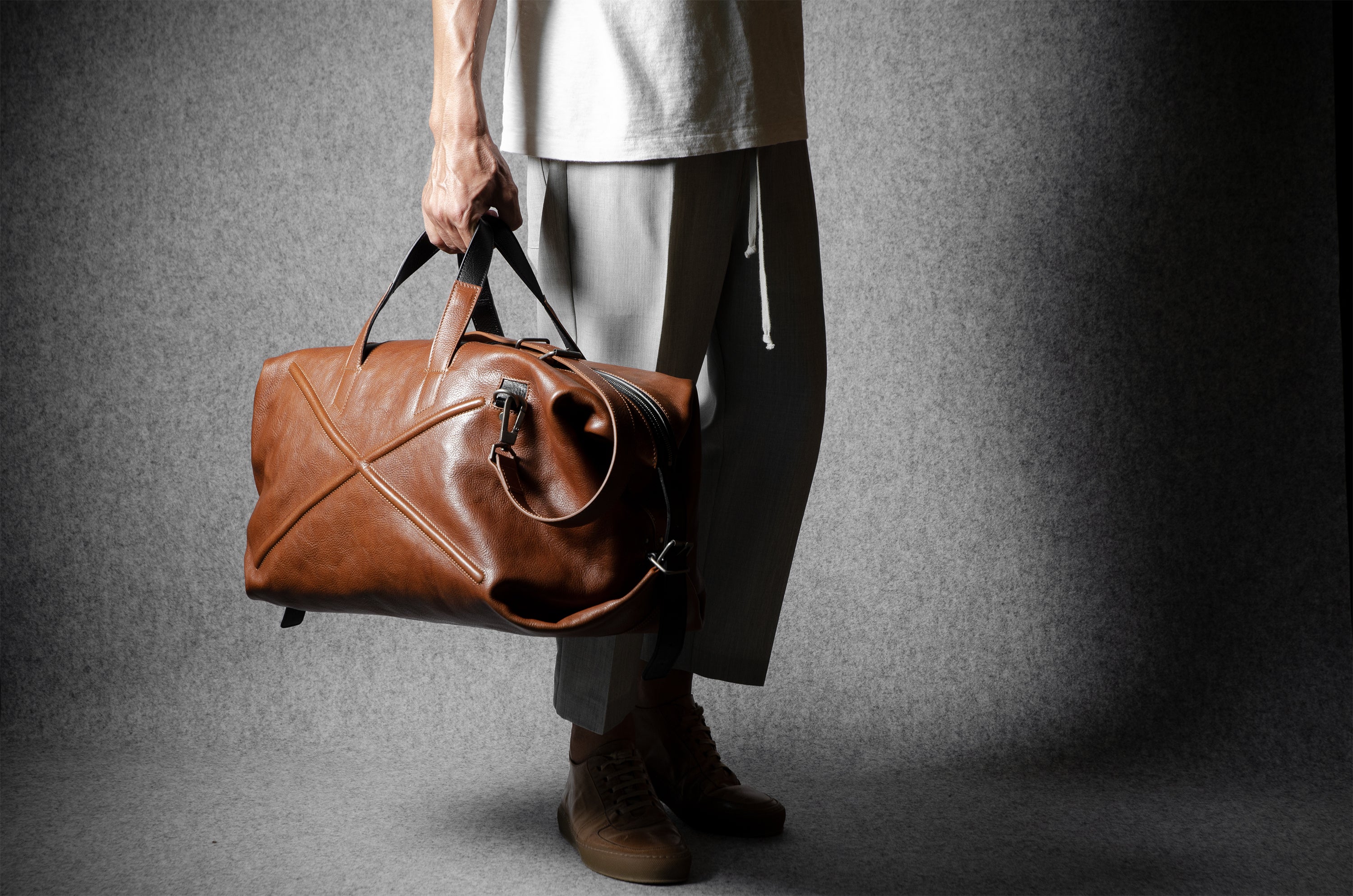 Person holding a brown leather duffel bag against a gray background