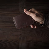 Brown leather card case held by hand on a dark wood background.