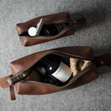 Two brown leather zippered pouches arranged on a gray textured surface, each open to show toiletry items inside.