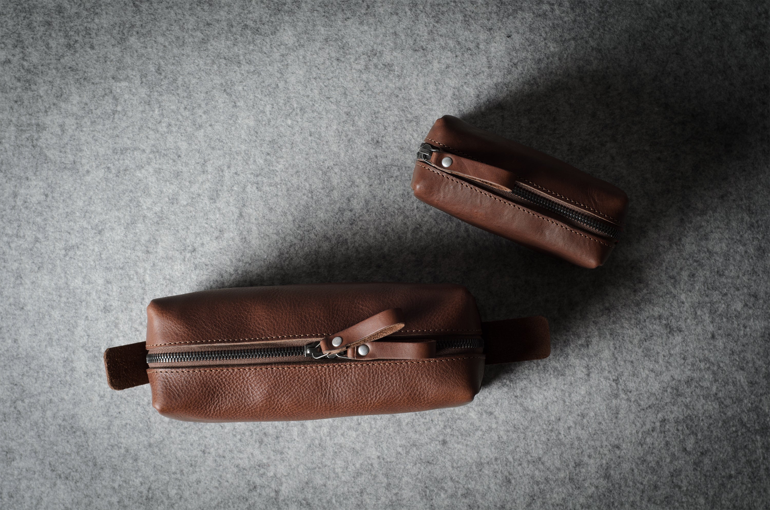 Two brown leather zippered dopp-kit pouches with leather pull tabs, displayed on a textured gray felt background.