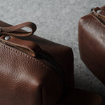 Close-up of a brown pebbled leather zip pouch with a leather zipper pull tab, shown on a textured gray surface with a second matching pouch partially visible in the background.