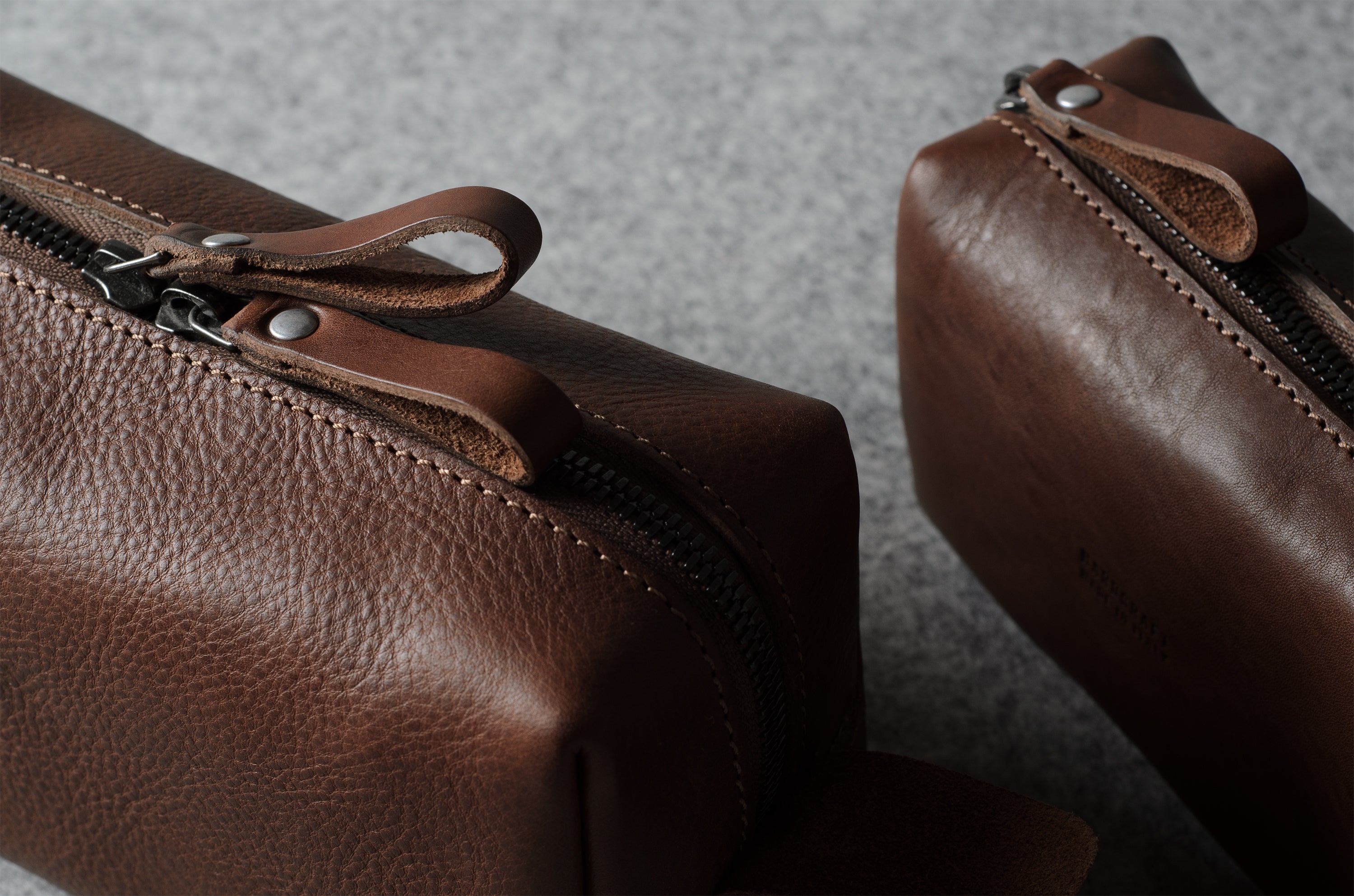 Close-up of a brown pebbled leather zip pouch with a leather zipper pull tab, shown on a textured gray surface with a second matching pouch partially visible in the background.
