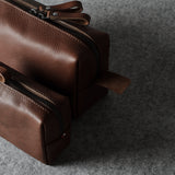 Two brown leather zippered pouches, one small and one larger, arranged diagonally on a textured gray felt surface.
