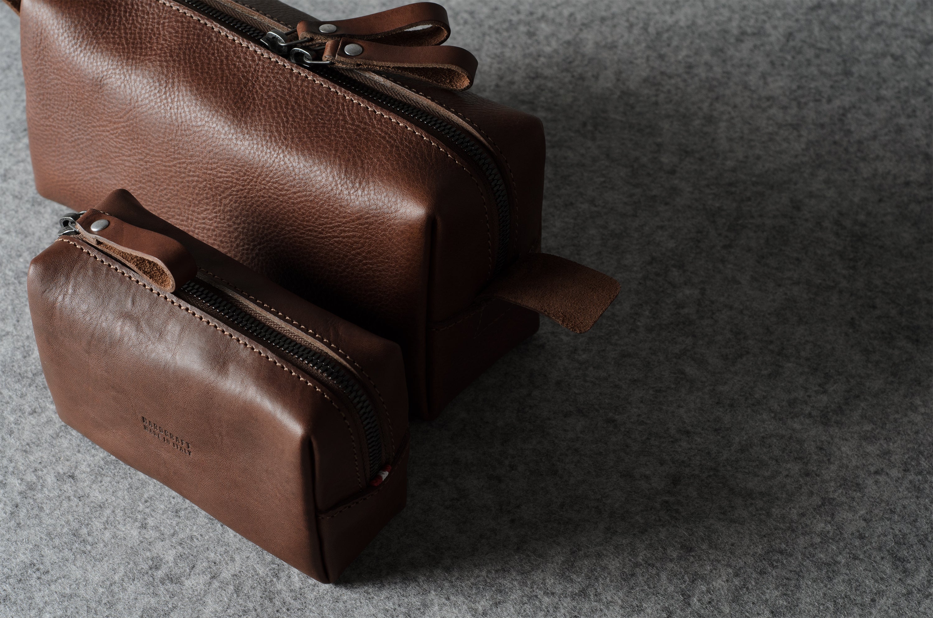 Two brown leather zippered pouches, one small and one larger, arranged diagonally on a textured gray felt surface.