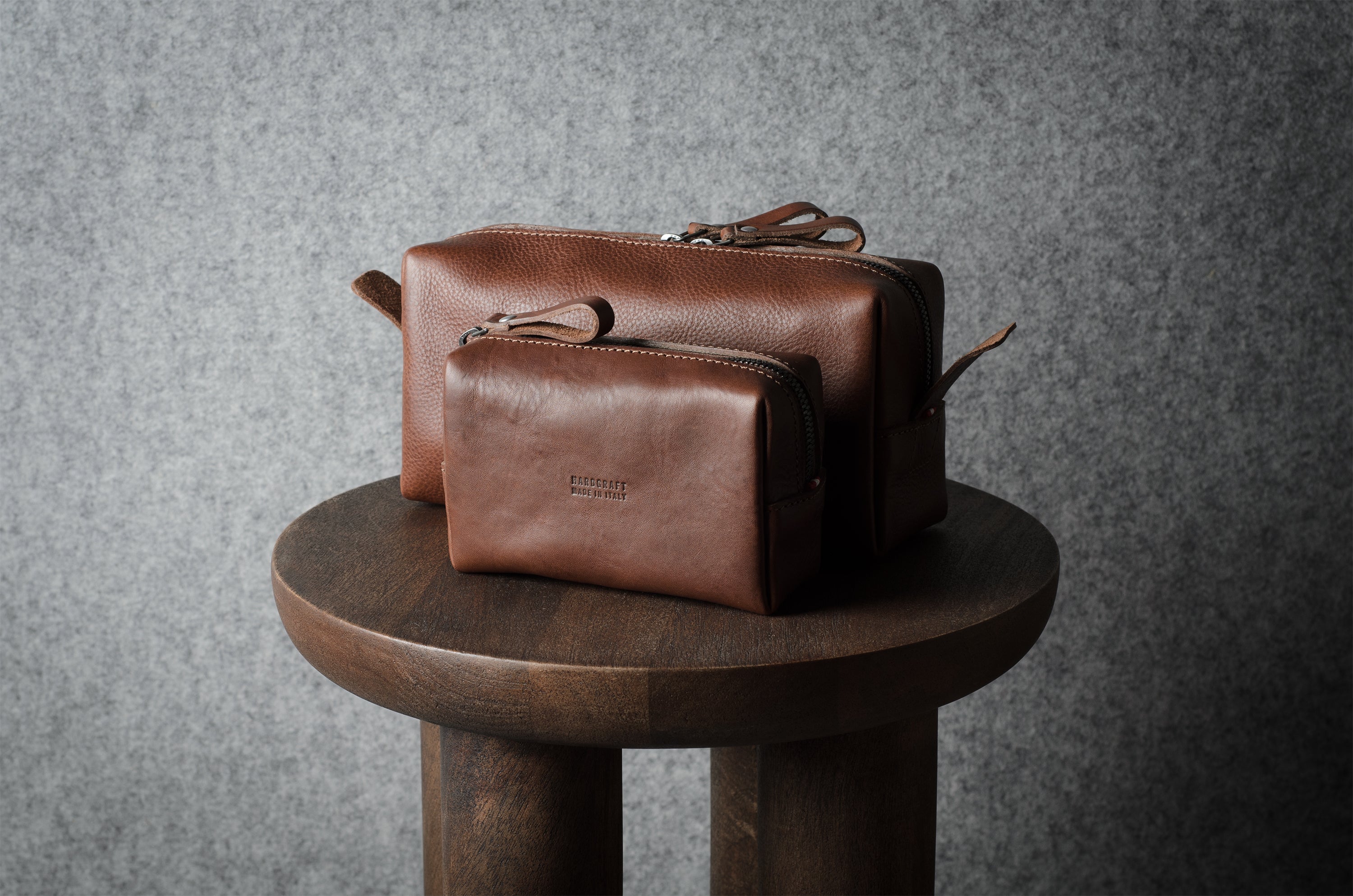 Two brown leather pouches of varying sizes stacked on a round wooden stool against a textured gray wall.