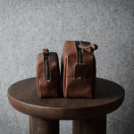 Two brown leather zippered pouches or small bags displayed side by side on a round wooden stool against a textured gray wall.