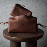 Two brown leather pouches stacked on a round wooden stool against a textured gray background.