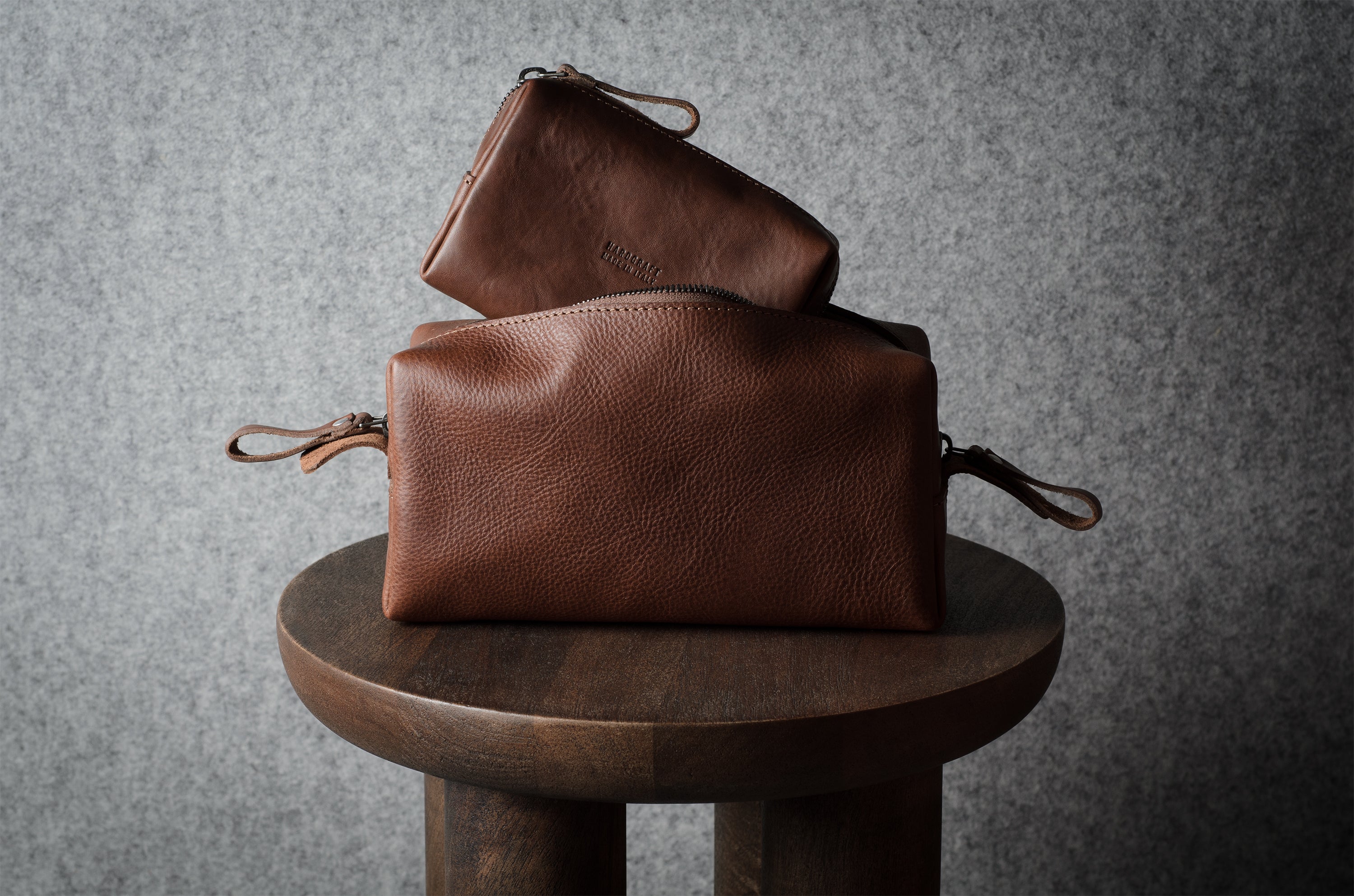 Two brown leather pouches stacked on a round wooden stool against a textured gray background.