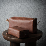 Two brown leather pouches, one larger and one smaller, displayed on a dark wooden stool against a textured gray background.