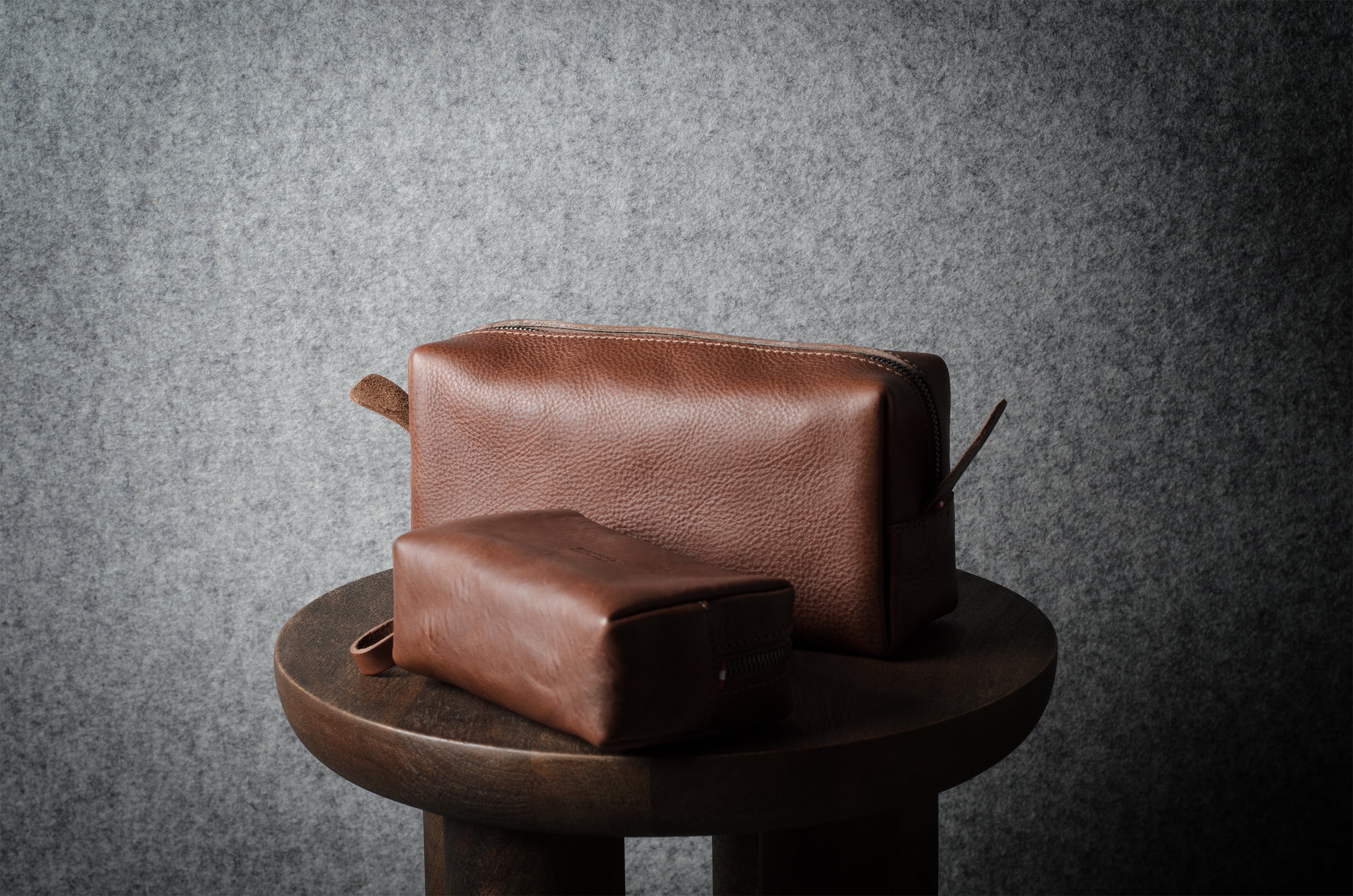 Two brown leather pouches, one larger and one smaller, displayed on a dark wooden stool against a textured gray background.