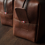 A close-up of a brown pebbled leather zippered pouch with a front pocket and a small red-and-white striped accent near the zipper, photographed on a dark wooden surface.