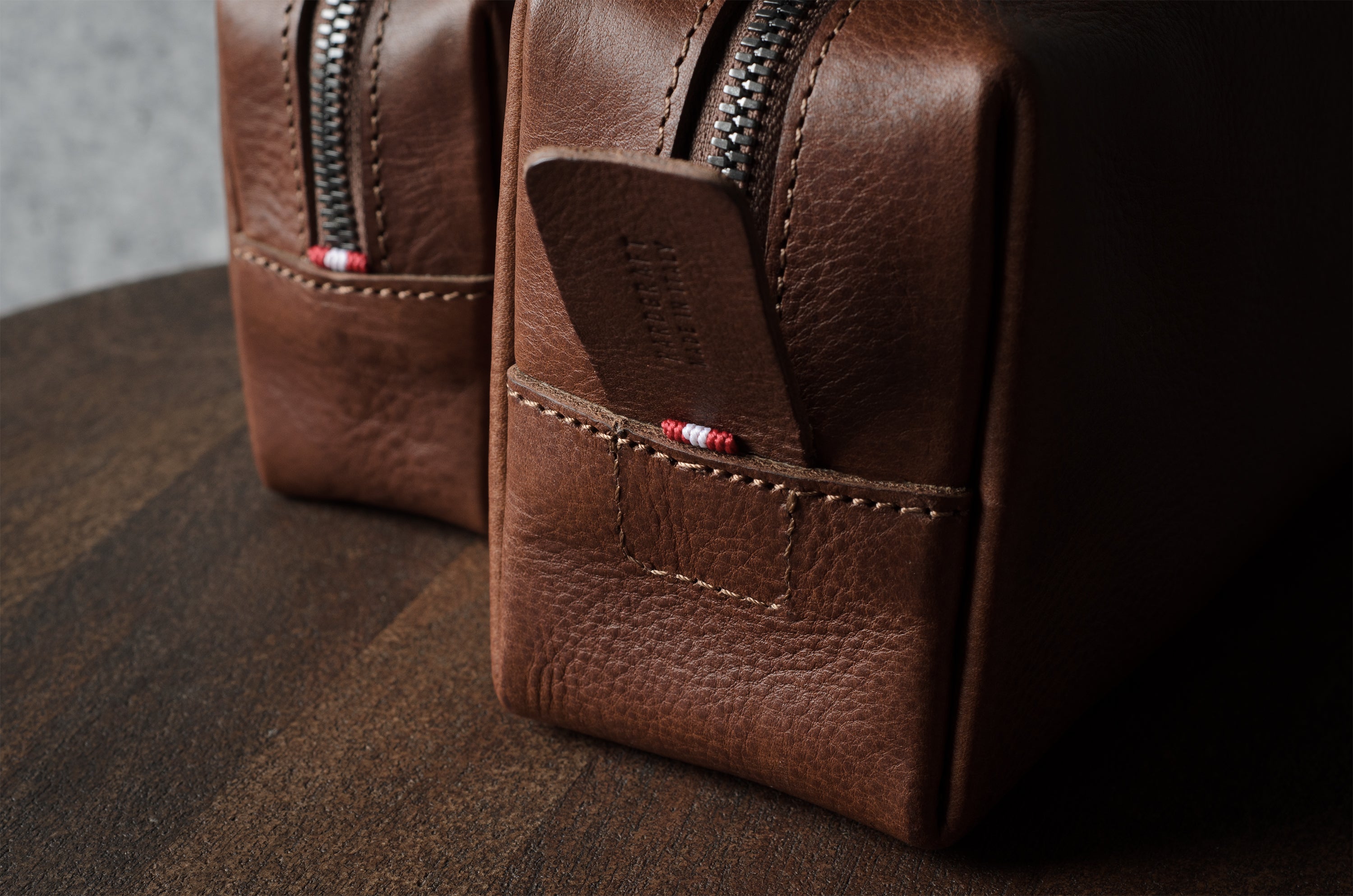 A close-up of a brown pebbled leather zippered pouch with a front pocket and a small red-and-white striped accent near the zipper, photographed on a dark wooden surface.