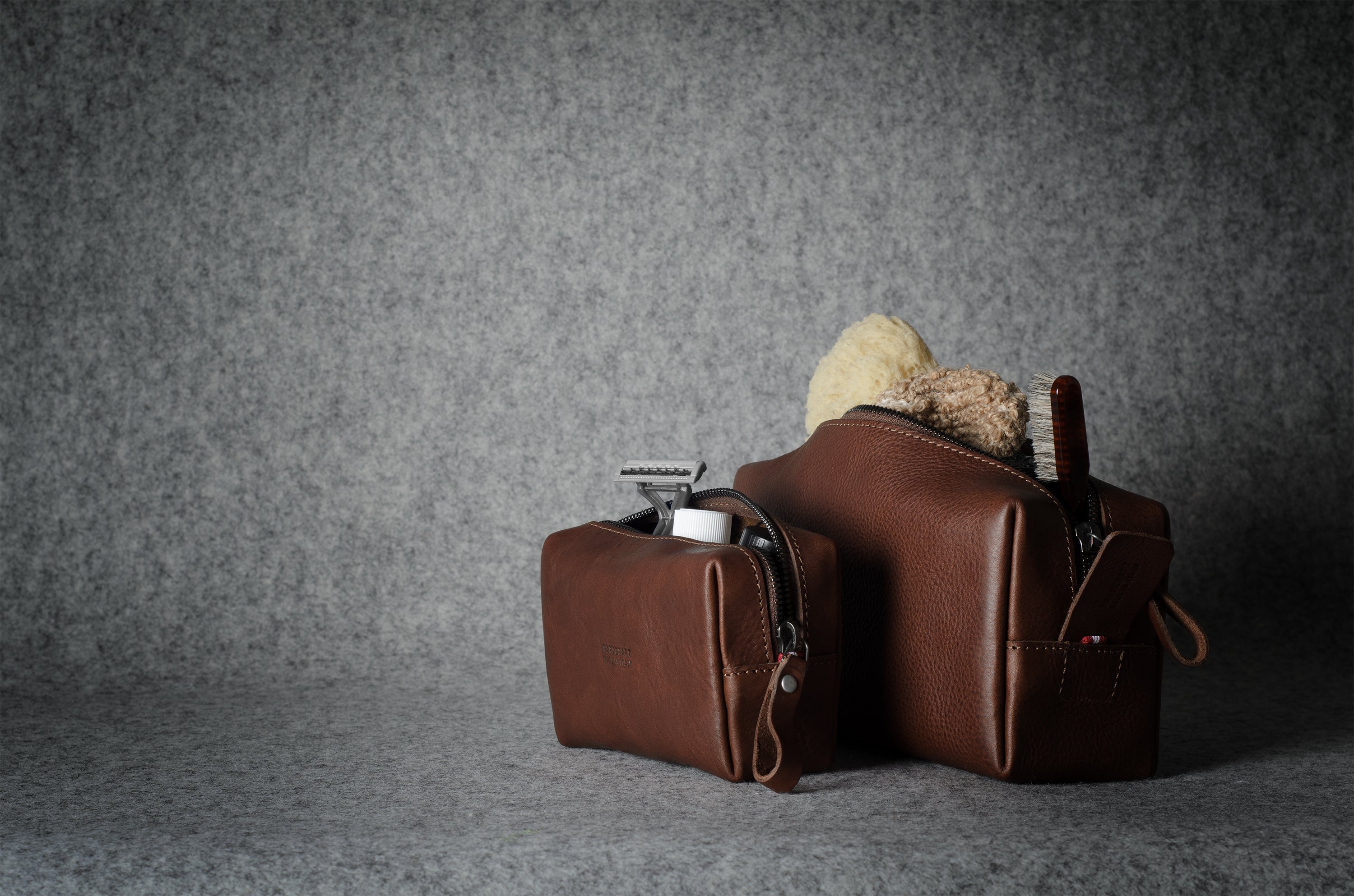 Two brown leather zippered pouches or small bags with loop handles, shown on a textured gray surface against a gray backdrop. One pouch is open, revealing a beige toothbrush and a small white item inside.