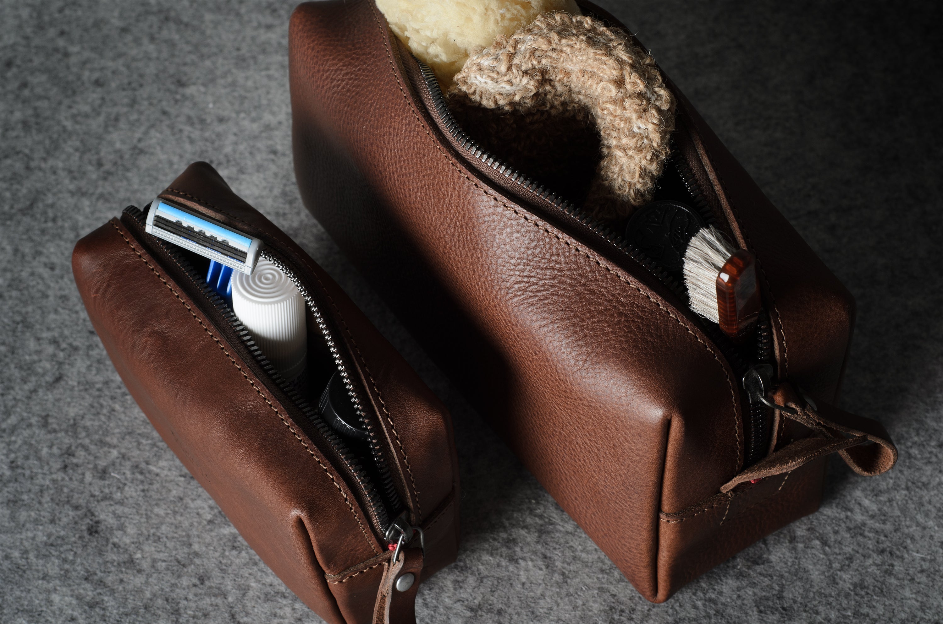 Two brown leather zippered toiletry bags on a gray textured surface, with one bag open to show a shaving razor and the other open to show a bath sponge.