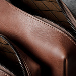 Close-up of a brown leather bag with a diagonal zipper opening, showing the pebbled leather texture and dark interior lining.