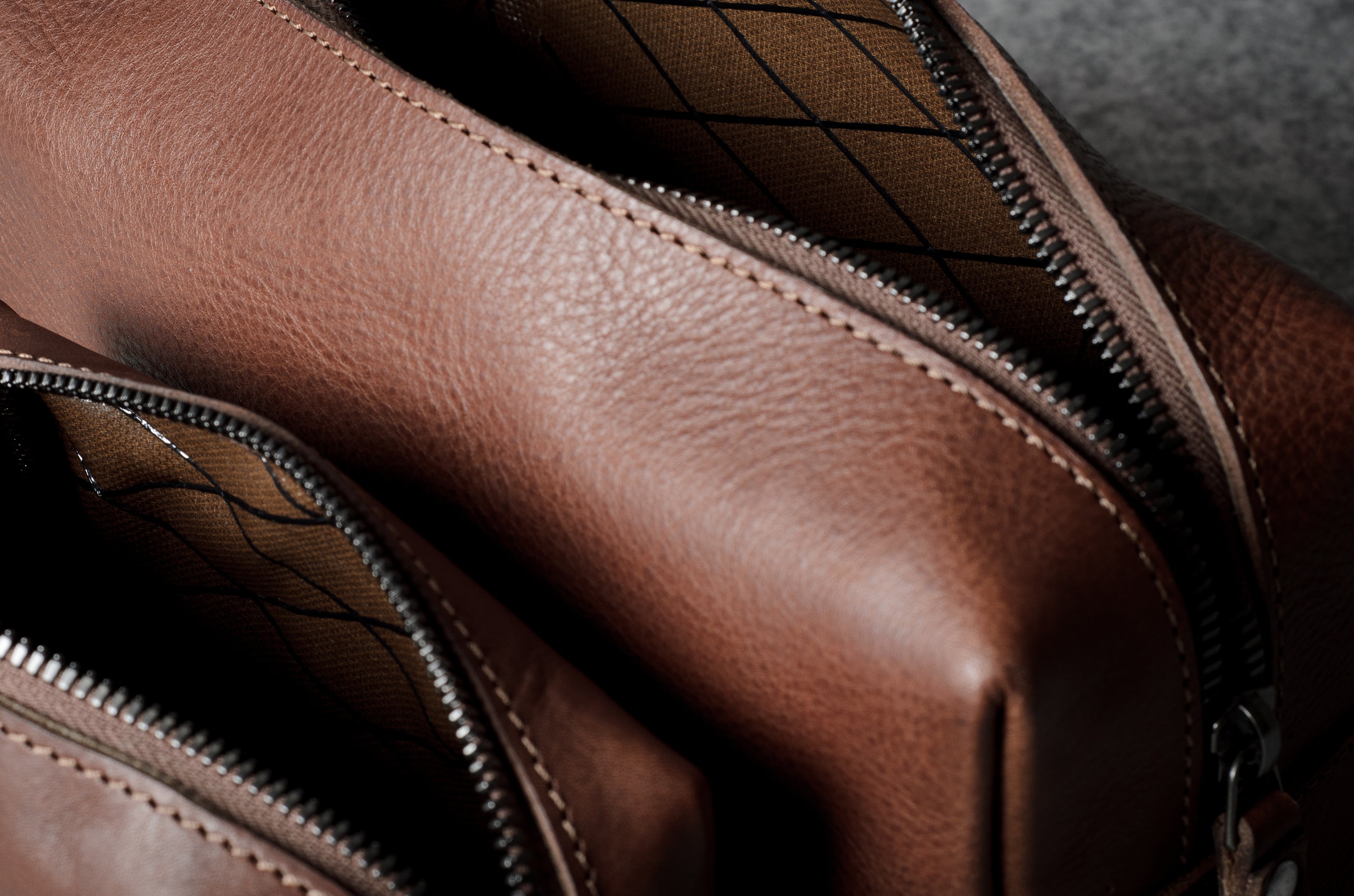 Close-up of a brown leather bag with a diagonal zipper opening, showing the pebbled leather texture and dark interior lining.