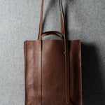A brown leather tote bag with long shoulder straps, hanging from a hook against a textured gray wall.
