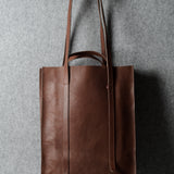 A brown leather tote bag with long shoulder straps, hanging from a hook against a textured gray wall.