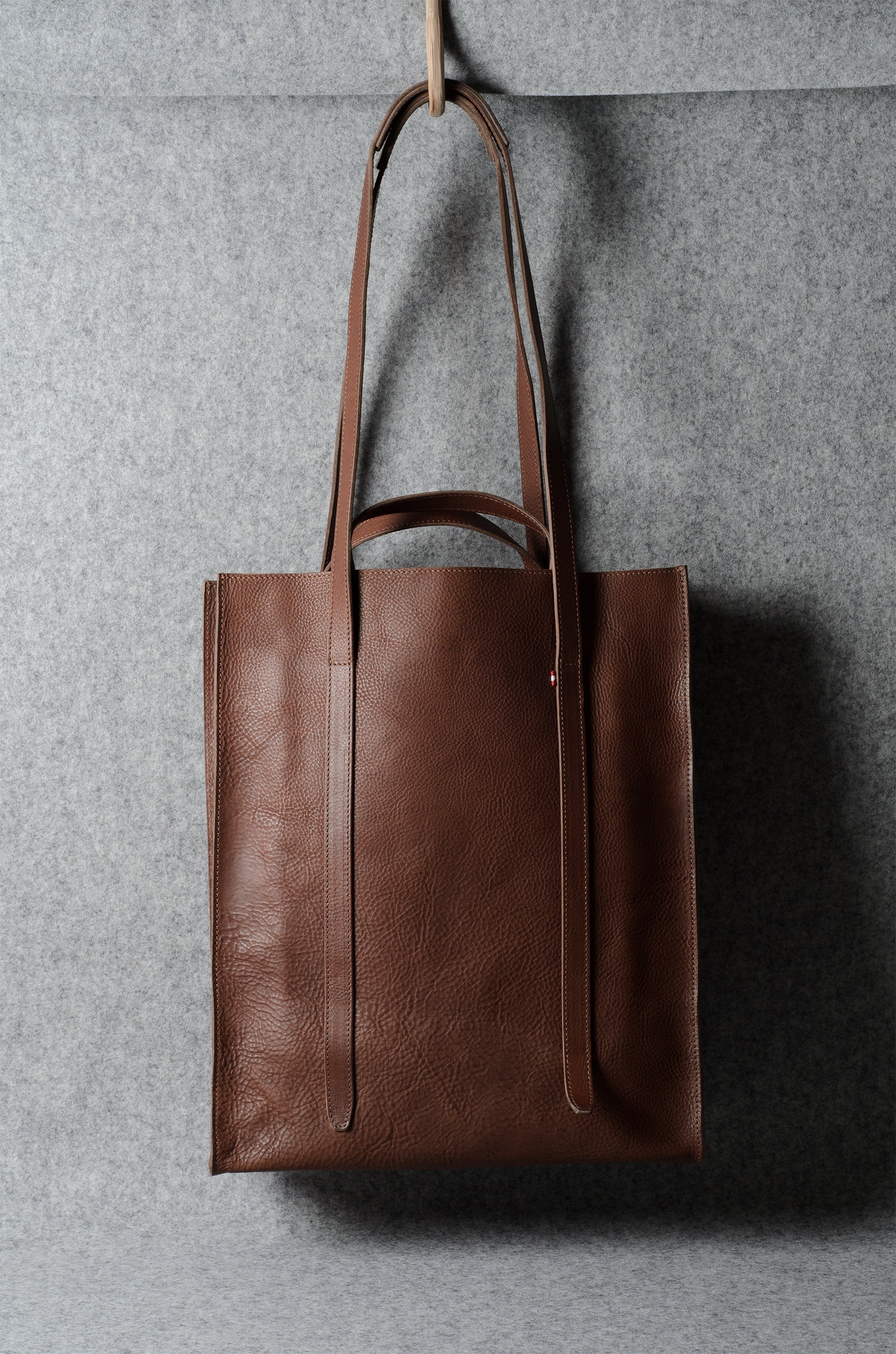 A brown leather tote bag with long shoulder straps, hanging from a hook against a textured gray wall.