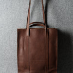 A brown leather tote bag hanging from a wooden hook against a textured gray wall.