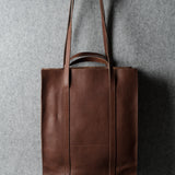 A brown leather tote bag hanging from a wooden hook against a textured gray wall.