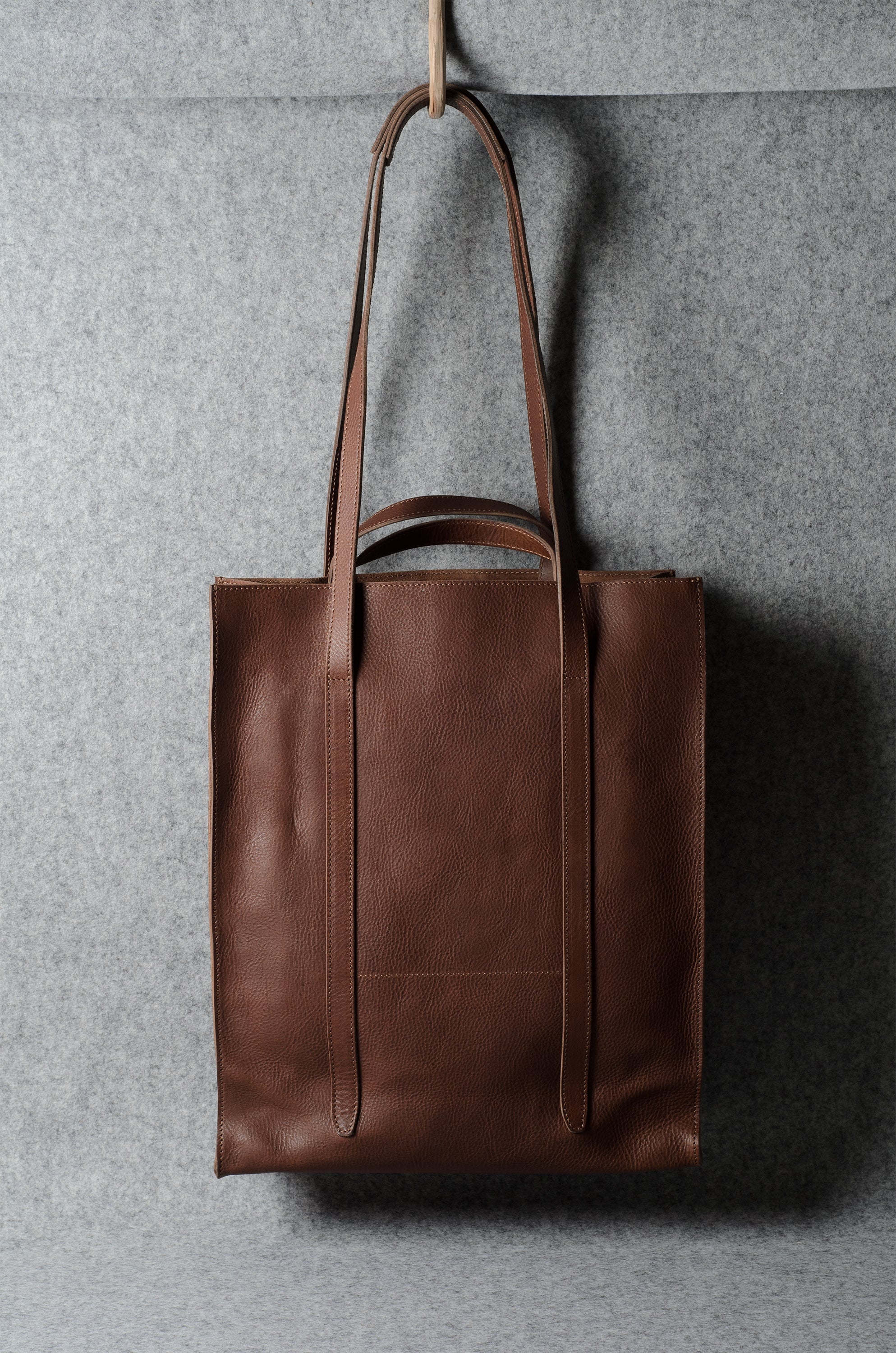 A brown leather tote bag hanging from a wooden hook against a textured gray wall.