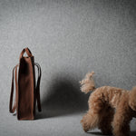 A brown leather tote bag stands on a gray studio floor next to a small fluffy brown dog, with the dog facing away from the camera.