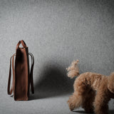 A brown leather tote bag stands on a gray studio floor next to a small fluffy brown dog, with the dog facing away from the camera.