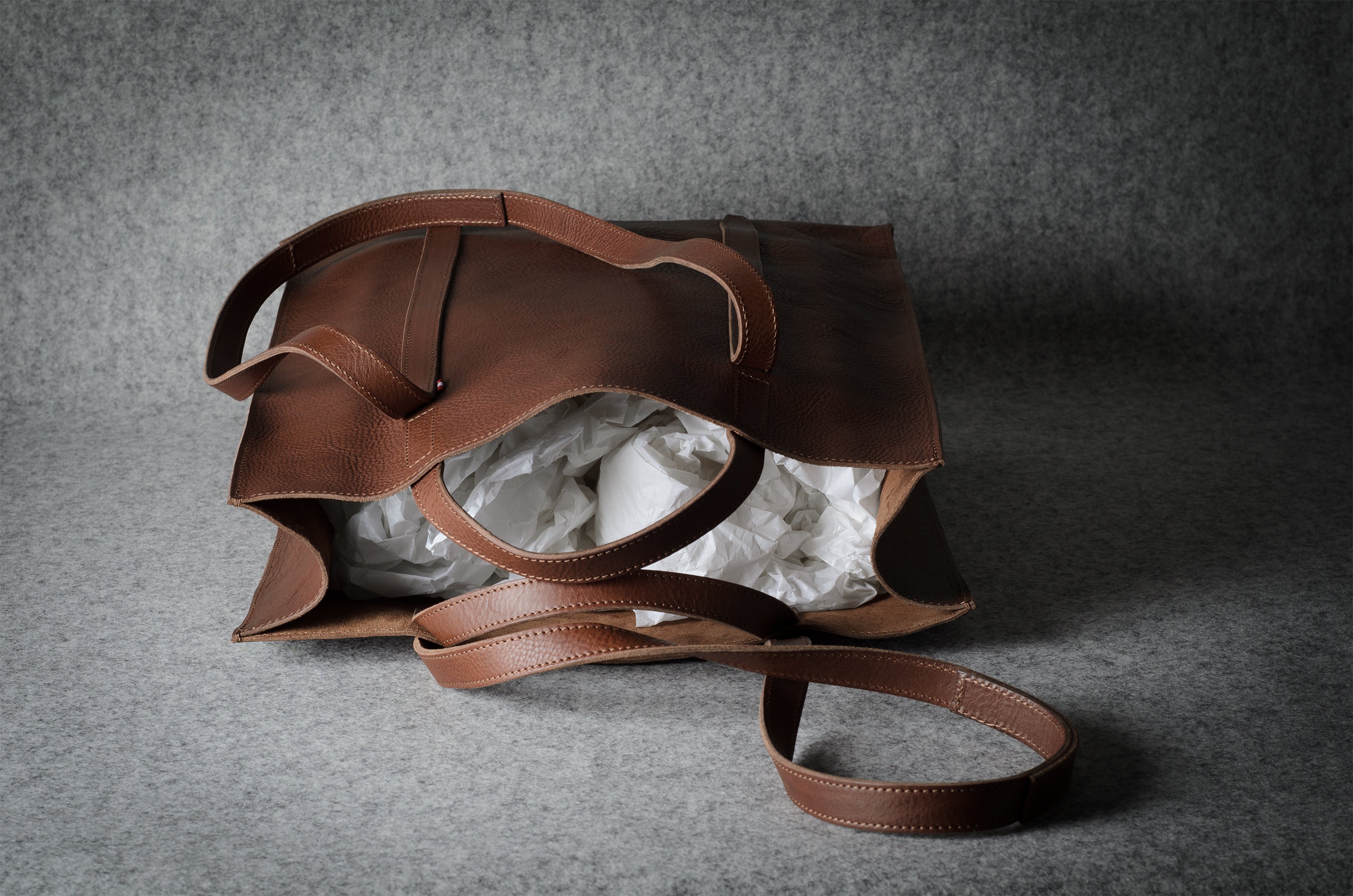 A brown leather tote bag with a matching shoulder strap, shown open with white tissue paper inside, set against a textured gray background.