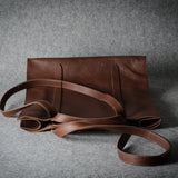 A brown leather tote bag with a matching shoulder strap, displayed on a textured gray background.