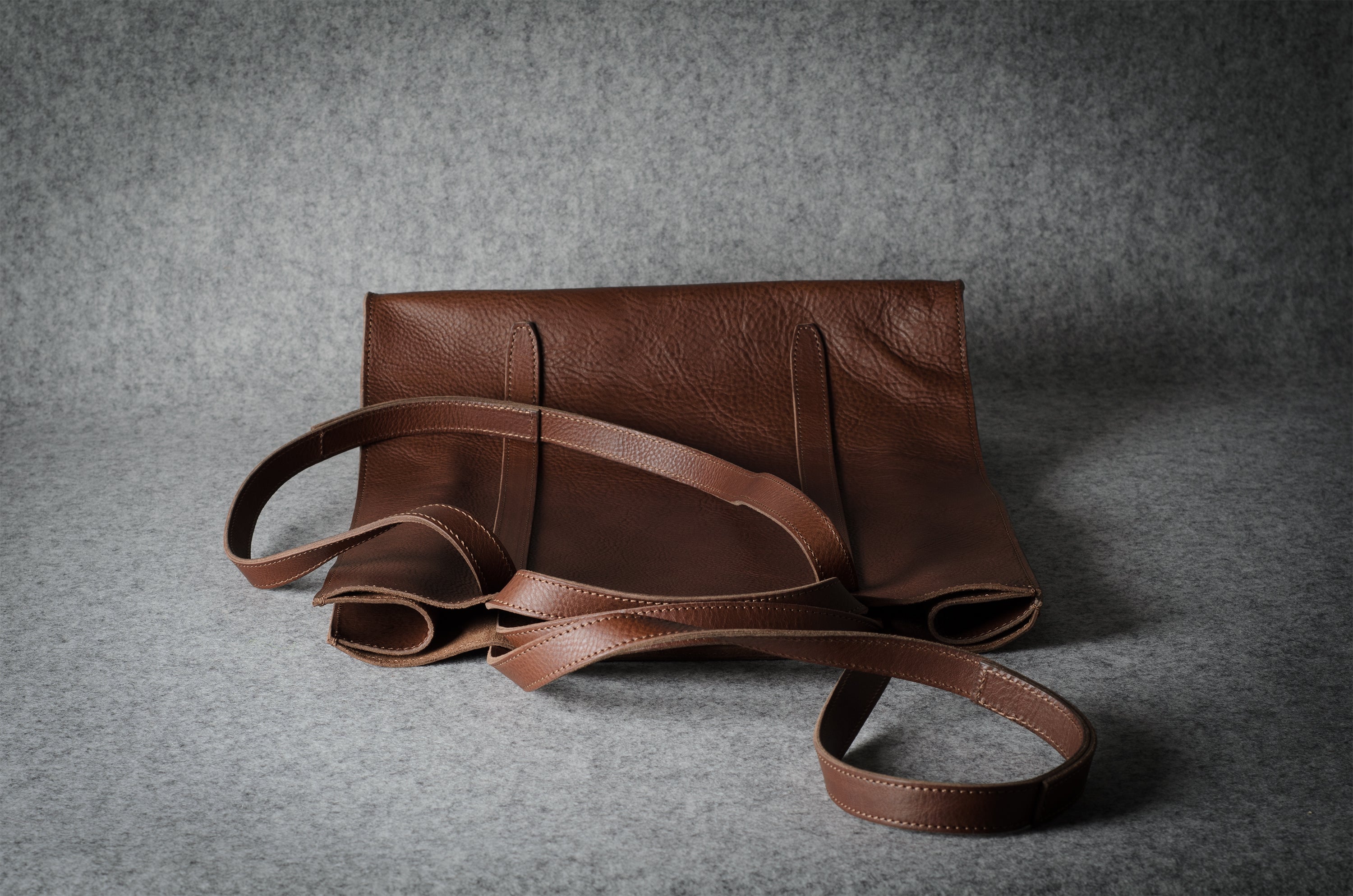 A brown leather tote bag with a matching shoulder strap, displayed on a textured gray background.